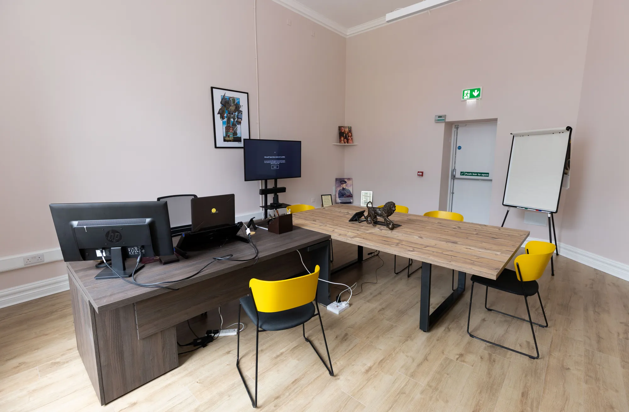 A meeting room with a desk, screen and oak table with yellow chairs